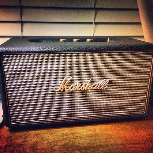 A Marshall home speaker with the brass script logo and textured vinyl front on a wooden desk in warm light