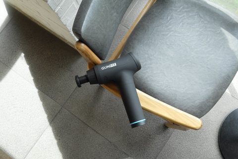 A small matte black percussion massage gun resting on a grey upholstered chair
