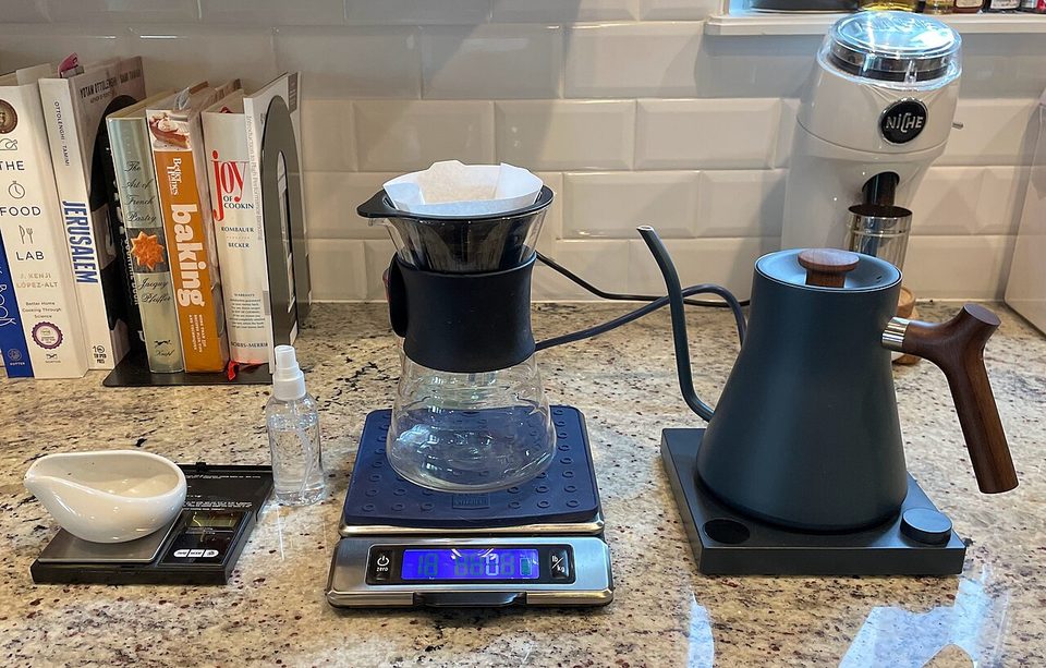 a matte black Fellow Stagg gooseneck kettle with a wood handle, beside a pour-over coffee dripper and a scale on a kitchen counter