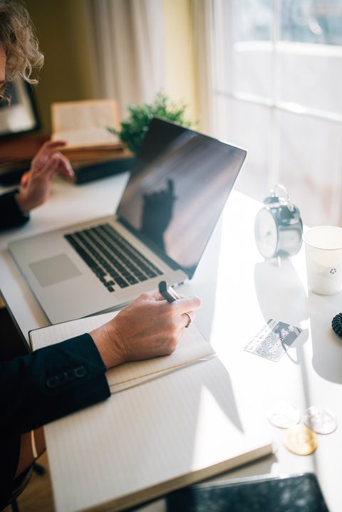 A person writing notes at a desk beside an open laptop