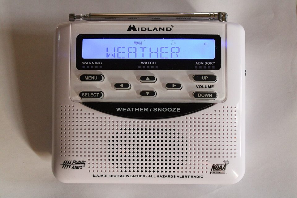 A Midland WR-120EZ NOAA weather radio on a table, a gray desktop box with a small text display, antenna up