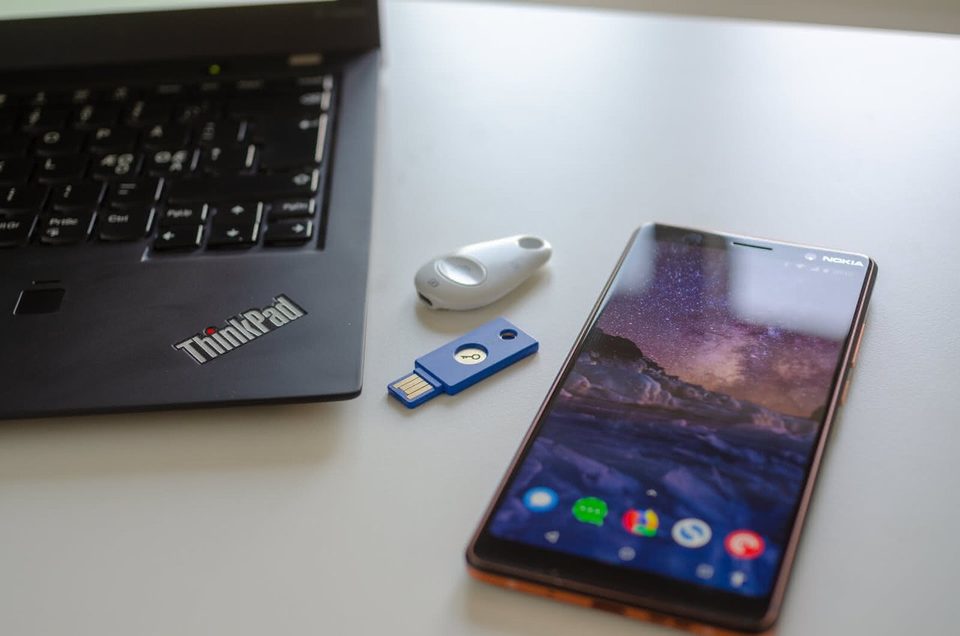 Two FIDO security keys from other makers, a white key fob and a blue USB key, between a ThinkPad and an Android phone, standing in for the Solo 2