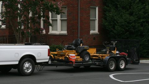 A pickup towing a trailer loaded with two commercial zero-turn mowers, parked outside a brick building