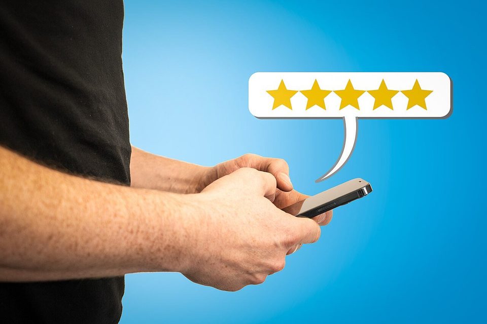 A person's hands typing on a smartphone below a five-star rating bubble, in a staged photo against a blue backdrop